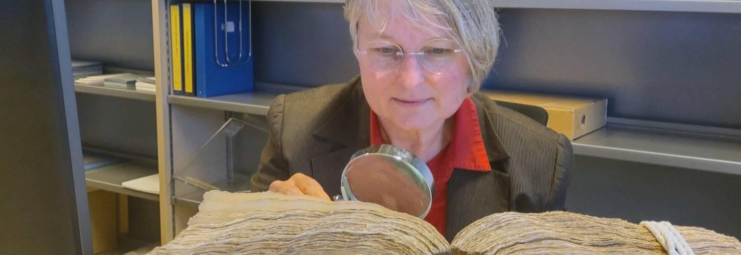 Ineke buigt zich met een loep over een groot oud dik boek dat voor haar ligt