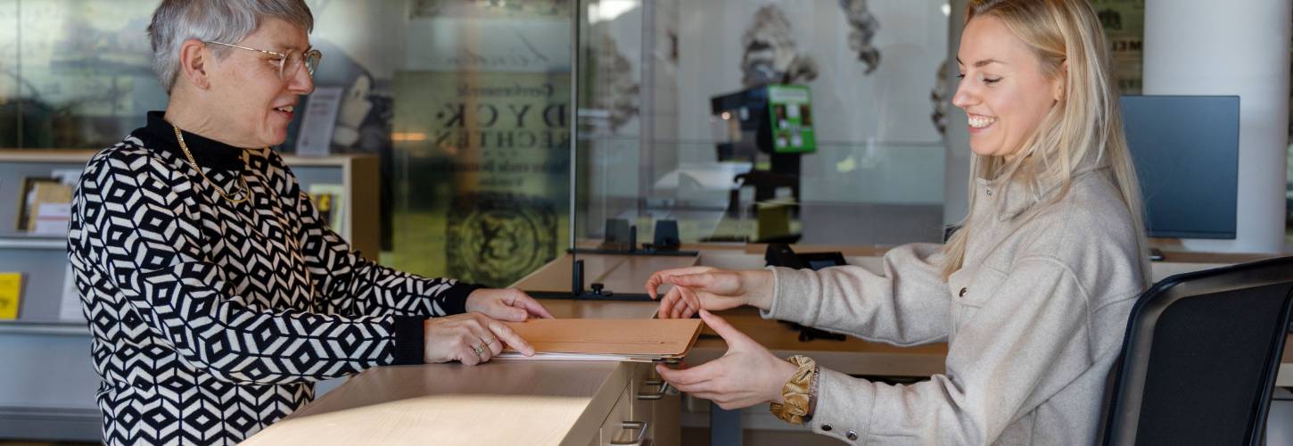 Medewerker RAR overhandigt archiefstukken aan studiezaalmedewerker aan de balie van de studiezaal
