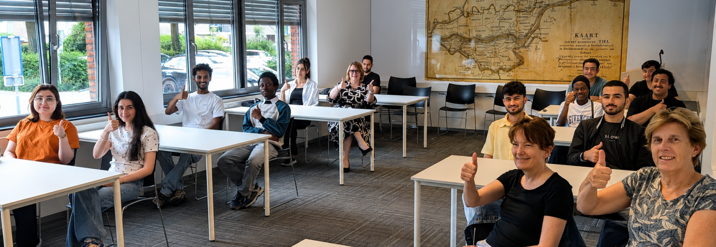 groep mensen twee aan twee aan tafels in een leslokaal met een kaart van Rivierenland op de muur achter hen, ze steken allemaal een duim op