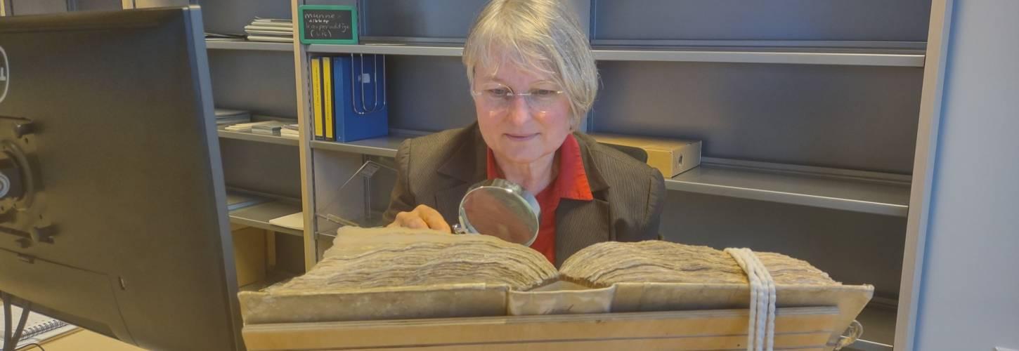 Vrijwilliger bekijkt een oud boek met een grote loep met op de voorgrond een computerscherm en op de achtergrond een open kast