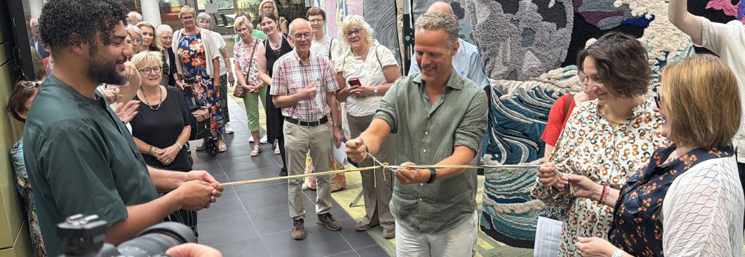 Een man knoopt draden aan elkaar die door een andere man en een vrouw worden vastgehouden terwijl anderen toekijken. Op de achtergrond is een groot wandkleed te zien.