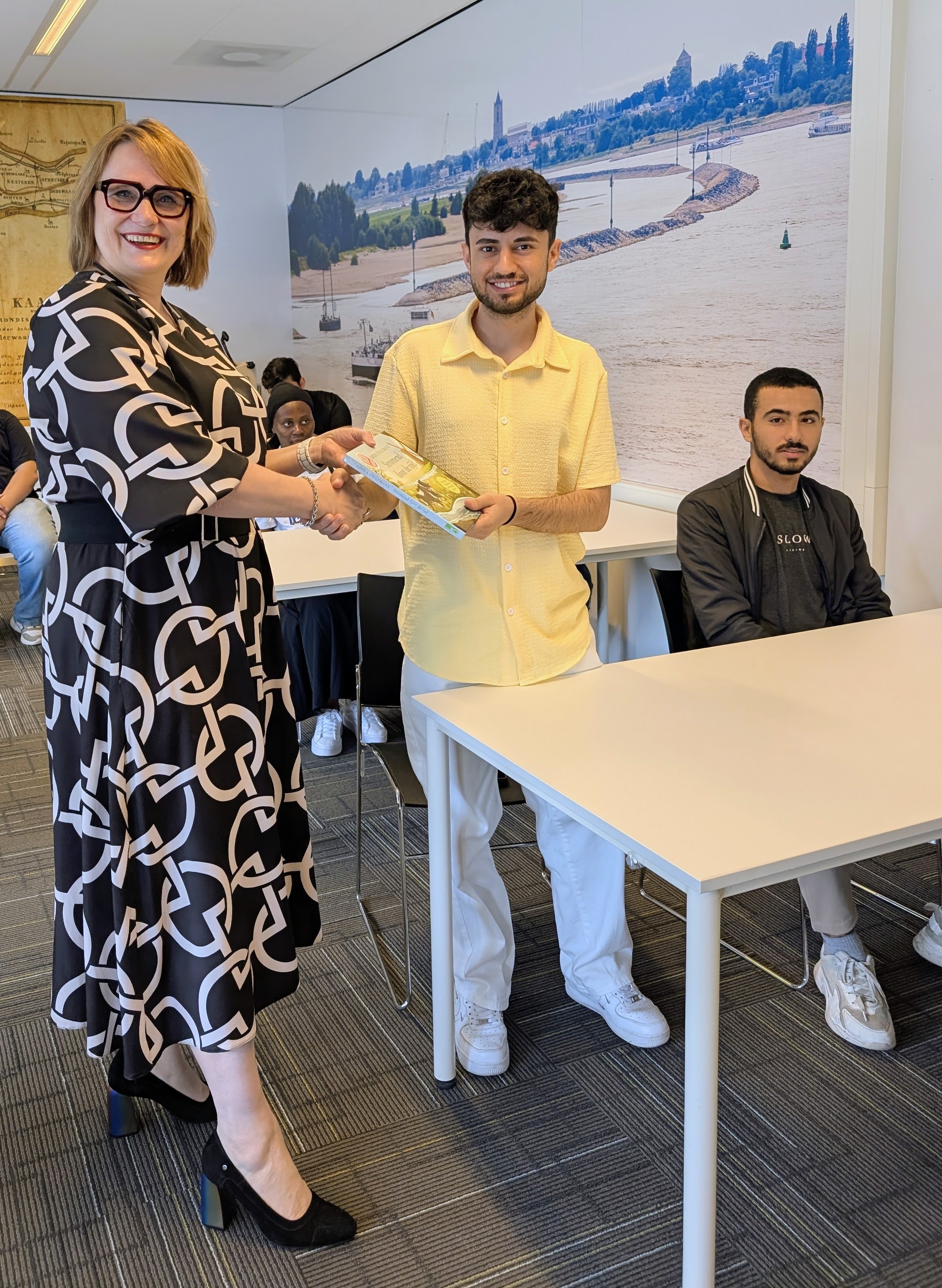 Een vrouw geeft een jonge man een hand en een boek in een leslokaal terwijl een zitende jongeman toekijkt