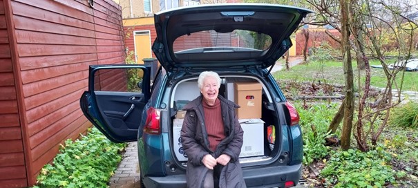 Marleen Kaptein zit op de rand van de achterkant van haar auto. De achterklep staat omhoog en achter haar staan kartonnen dozen.